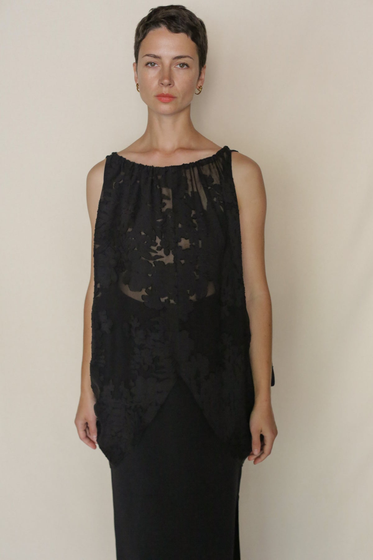 A person with short dark hair wears the Uragiri Butterfly, a sleeveless black dress with a sheer floral overlay, standing against a plain light background and looking directly at the camera with a neutral expression.