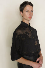 Load image into Gallery viewer, A person with short dark hair and gold earrings wears a sheer black Work Shirt | Silk Uragiri Black with floral patterns and gold buttons, standing against a plain light background and looking slightly to the side.