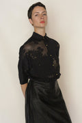 A person with short dark hair wears a sheer black embroidered Uragiri Shirt and a shiny black Lan Jaenicke skirt, standing against a plain light background, looking confidently at the camera.