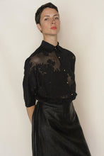 Load image into Gallery viewer, A woman with short dark hair confidently wears Lan Jaenicke's Work Shirt | Silk Uragiri Black, a sheer blouse with embroidered floral patterns, paired with a shiny black skirt, standing against a plain light background.