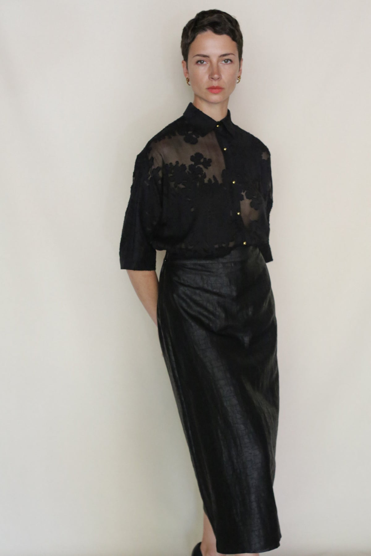 A person with short hair stands against a plain background, wearing the Uragiri Shirt paired with a high-waisted, long black skirt. Their hands are behind their back and they have a neutral expression.