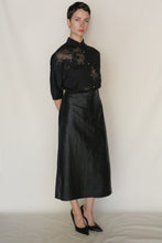 Load image into Gallery viewer, A person stands against a plain light background, wearing a sheer black floral blouse, the Futo Skirt | Xiang Yun Silk Earth Mark with a subtle crackled surface, and black pointed heels. They have short hair and a neutral expression.
