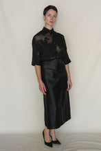 Load image into Gallery viewer, A person stands against a plain background wearing a sheer black floral blouse, black pointed-toe heels, and the Futo Skirt | Xiang Yun Silk Earth Mark—a long handmade skirt with a crackled surface. They have short dark hair and a neutral expression.