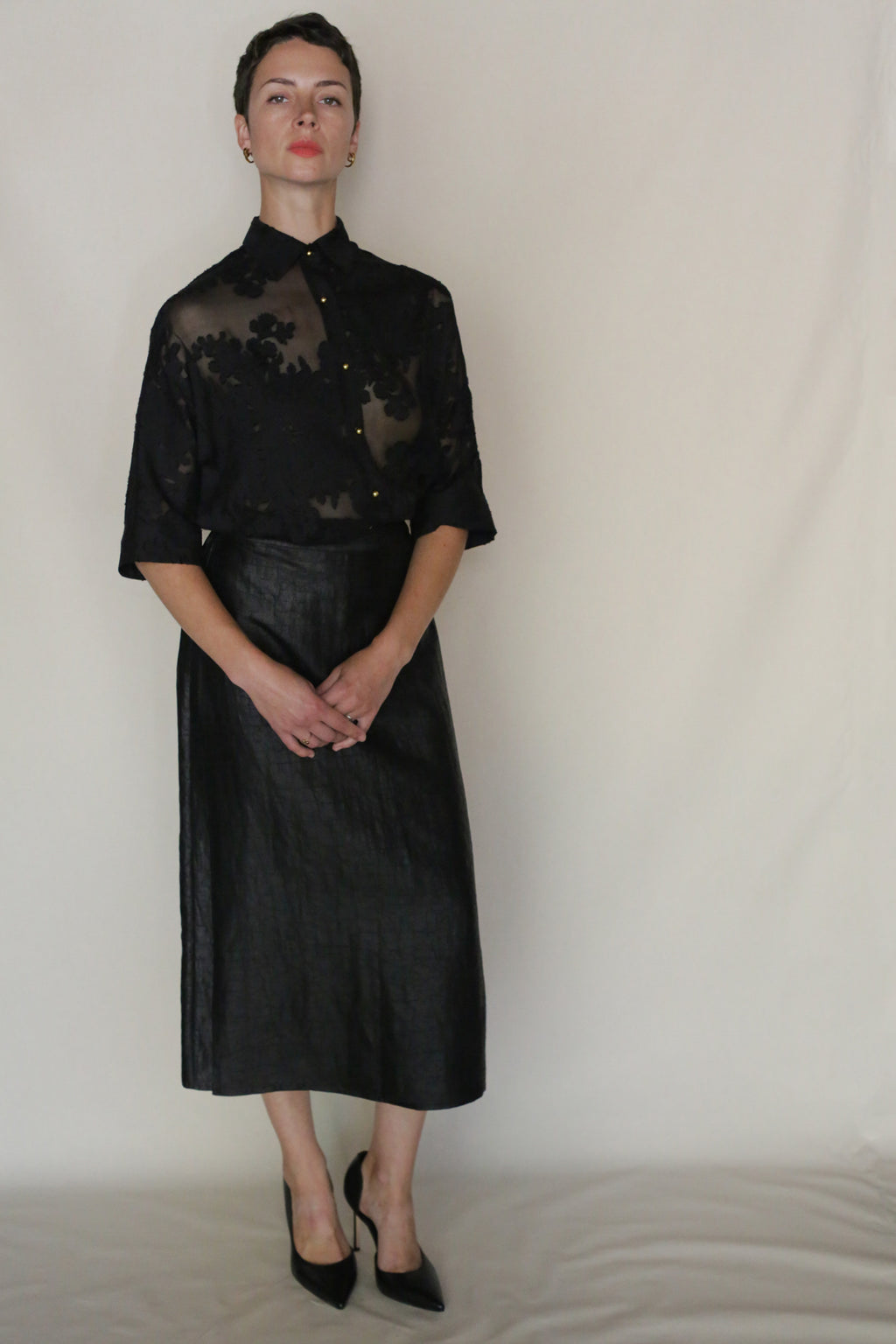 A person stands against a plain light background wearing a sheer black floral blouse, black heels, and the handmade Earth Mark Futo Skirt in a wrap silhouette. Their hands are folded in front as they look directly at the camera.