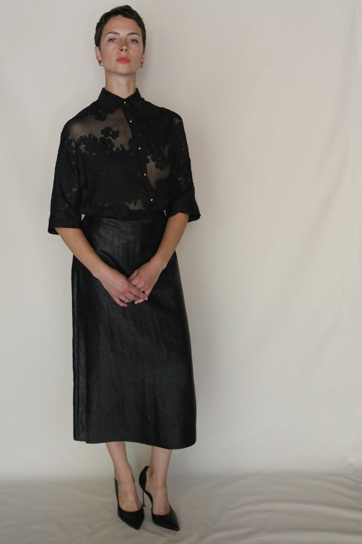 A person stands against a plain light background wearing a sheer black floral blouse, black heels, and the handmade Earth Mark Futo Skirt in a wrap silhouette. Their hands are folded in front as they look directly at the camera.