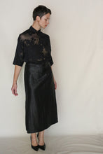 Load image into Gallery viewer, A person in a sheer black floral blouse and the Futo Skirt | Xiang Yun Silk Earth Mark stands against a light background. With short hair, gold earrings, and black heels, they look down, one arm relaxed by their side.