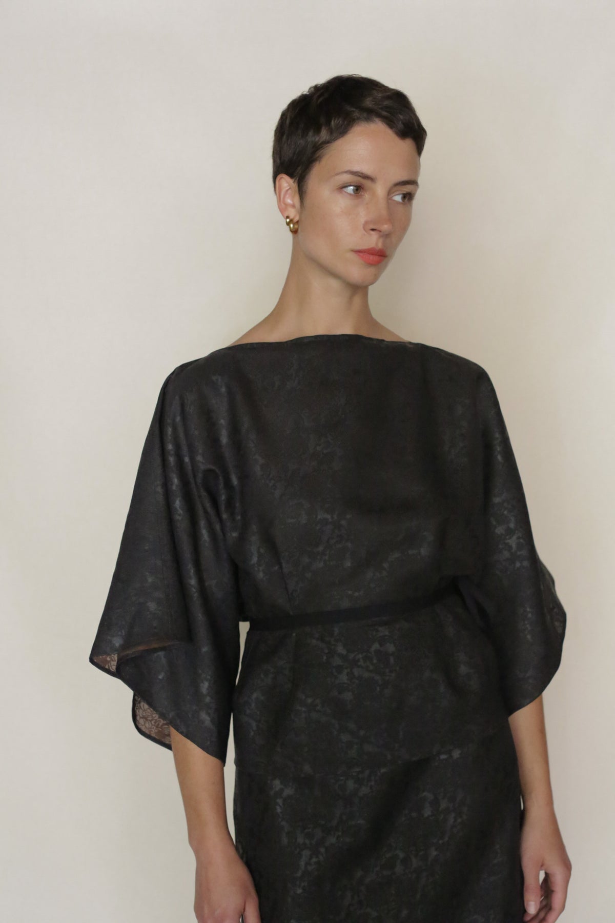 A woman with short dark hair wears the Endless Bloom Kimono Top—black, loose-fitting with wide sleeves—standing against a plain light background and gazing to the side with a neutral expression.