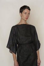 Load image into Gallery viewer, A woman with short dark hair and gold earrings wears a loose, dark Kimono Top Reversible | Xiang Yun Silk Endless Bloom with wide sleeves, standing against a plain light background, gazing to the side with a neutral expression.