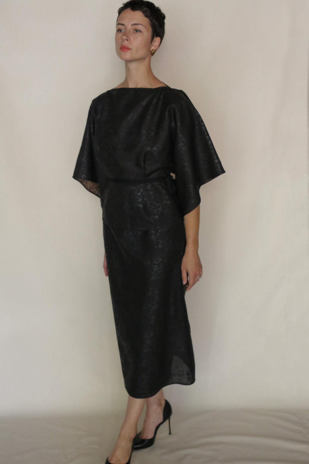 A woman in a loose-fitting, shiny black Endless Bloom Kimono Top with wide sleeves stands against a plain light background, wearing black heels. She gazes slightly upward with a neutral expression, hands relaxed at her sides.