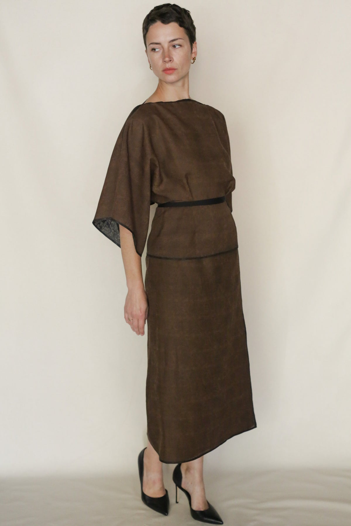 A woman stands against a plain background wearing the Endless Bloom Kimono Top, a loose-fitting brown silk piece with elbow-length sleeves, paired with a black belt and heels. She looks to the side with her hands at her sides.