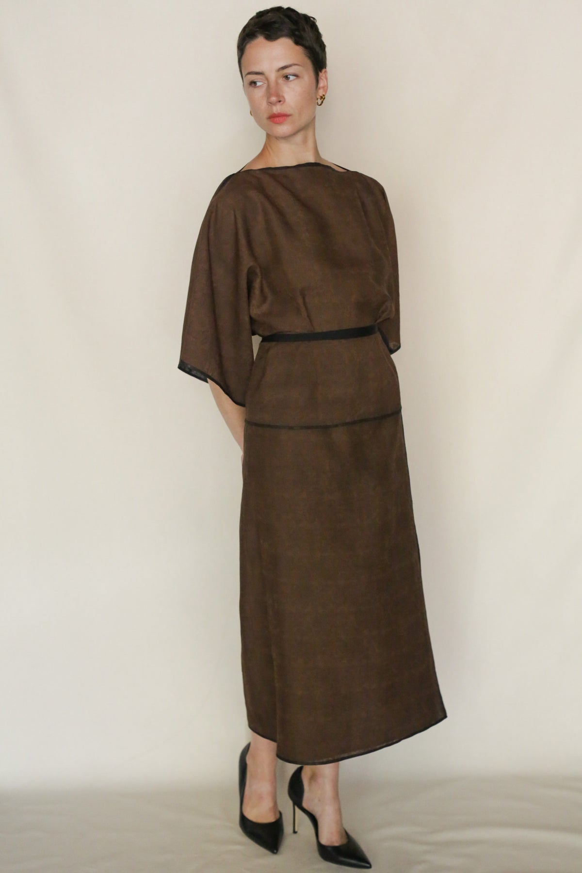 A woman stands against a light background, looking to the side, wearing the Endless Bloom Futo Skirt with black pointed-toe heels and a loose-fitting brown silk jacquard dress featuring wide sleeves and black trim.