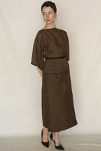 Load image into Gallery viewer, A person wearing the Reversible Futo Skirt in Xiang Yun Silk Endless Bloom, a loose two-piece brown outfit with wide sleeves and subtle black trim, stands against a cream background looking thoughtfully to the side in black heels.
