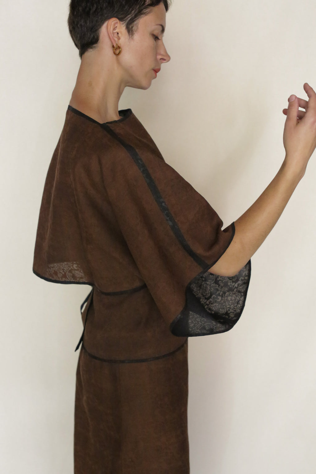 A person with short dark hair, wearing the Endless Bloom Kimono Top with wide sleeves and black trim, poses in profile against a plain light background, arm slightly raised and gaze downward.