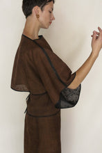 Load image into Gallery viewer, A person with short dark hair stands in profile against a plain background, raising one hand gracefully while wearing the Kimono Top Reversible | Xiang Yun Silk Endless Bloom in brown with black trim, looking down.