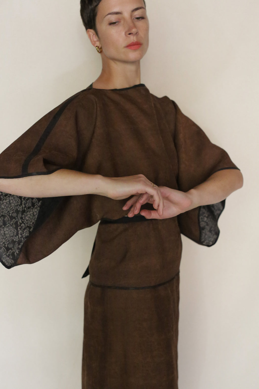Wearing the Endless Bloom Kimono Top in brown with wide sleeves, a person gracefully poses against a plain light background, arms extended and eyes gently closed.