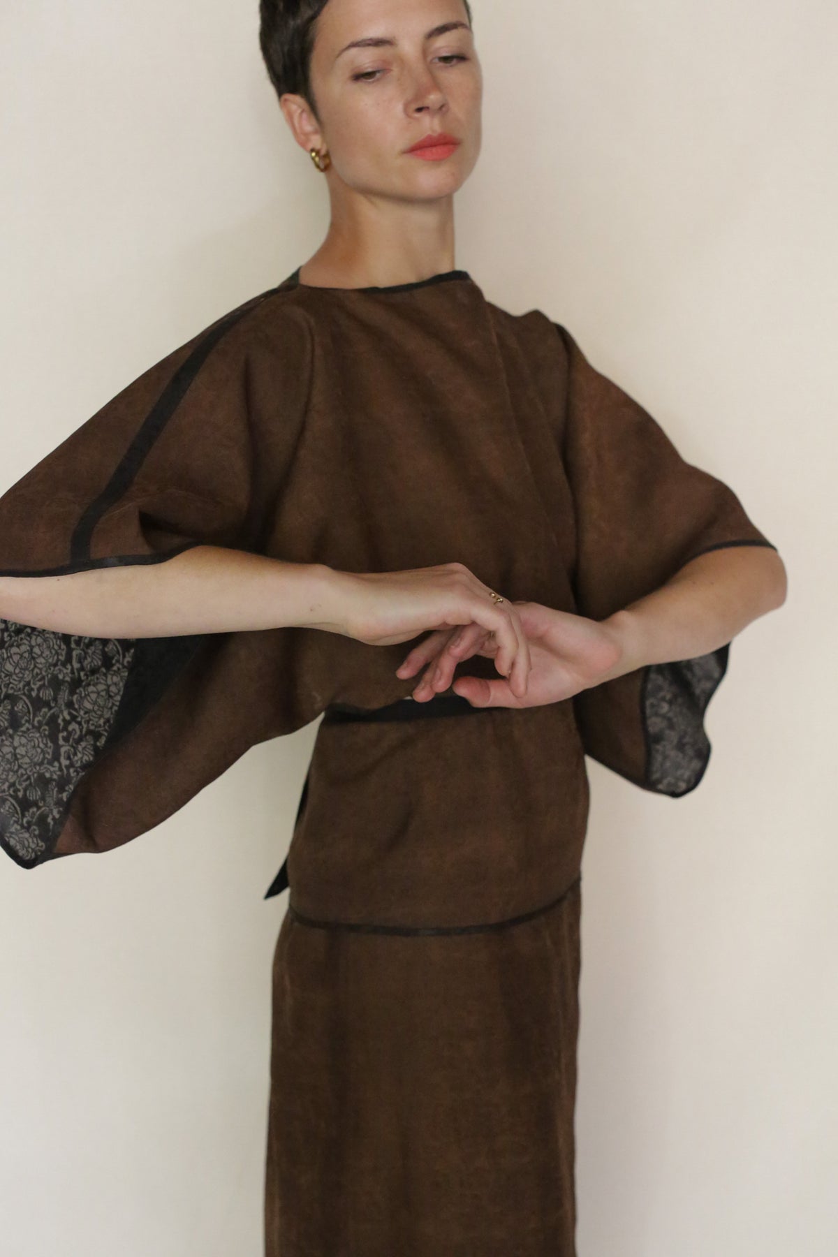 Wearing the Endless Bloom Kimono Top in brown with wide sleeves, a person gracefully poses against a plain light background, arms extended and eyes gently closed.