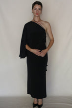 Load image into Gallery viewer, A person with short hair and minimal accessories stands against a plain background wearing the Eurie Blouse in 2 Ply Silk Crepe Black, styled as a one-shoulder dress with black heels, hands clasped in front.