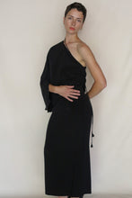 Load image into Gallery viewer, A person with short hair poses against a plain background, wearing the Eurie Blouse in 2 Ply Silk Crepe Black—styled as a one-shoulder, ankle-length dress with asymmetrical sleeve and tassel details.
