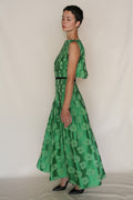 A woman with short hair wears the Hand Pleated Column Dress Hidden Peony, a sleeveless, floor-length silk-cotton jacquard dress with a floral pattern, paired with black heels, standing sideways against a plain light background.