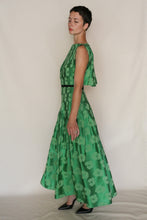 Load image into Gallery viewer, A woman stands in profile wearing the Hand Pleated Column Dress Hidden Peony Emerald, a sleeveless long silk-cotton jacquard dress with a floral pattern, paired with black heels against a plain light background.