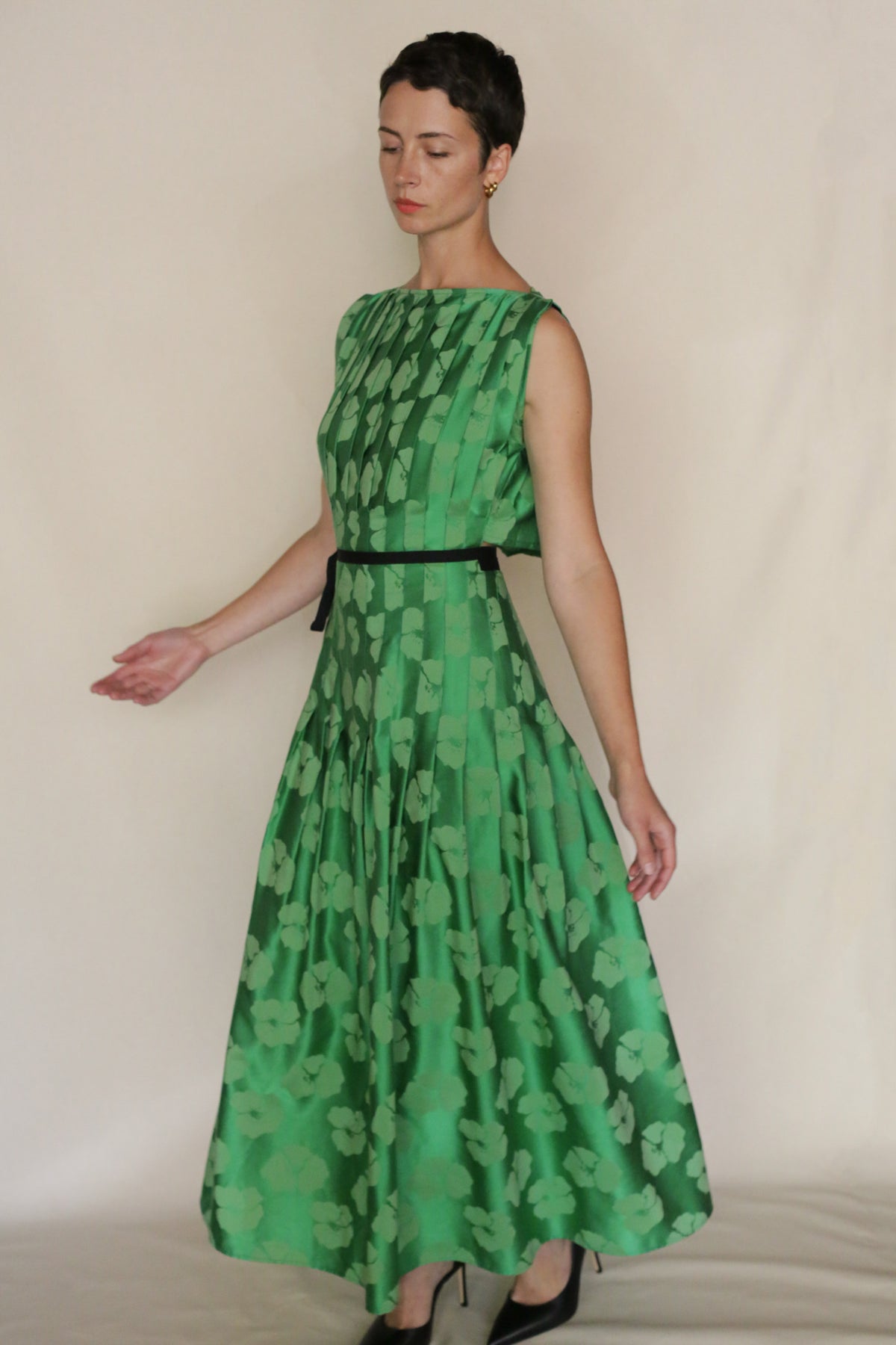 A person with short dark hair wears the Hand Pleated Column Dress Hidden Peony, an ankle-length floral jacquard dress with a black belt, standing against a plain light background, looking downward with arms slightly extended.