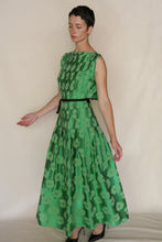Load image into Gallery viewer, A woman with short dark hair models the Hand Pleated Column Dress Hidden Peony Emerald—sleeveless, green, floral jacquard with hand-stitched pleats and a black belt. She stands sideways, eyes closed, arms gently outstretched against a plain backdrop.