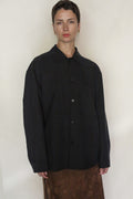 A person with short hair wears the Earth Mark Men's Shirt, loose-fitting and black, paired with a subtly patterned brown skirt, standing against a plain light background in a look inspired by natural plant dye aesthetics.