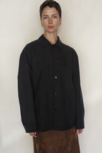 Load image into Gallery viewer, A person with short hair wears the Men's Shirt | Xiang Yun Silk Earth Mark, a loose-fitting, dark button-up shirt, paired with a brown Xiang Yun Silk skirt, standing against a plain light background.