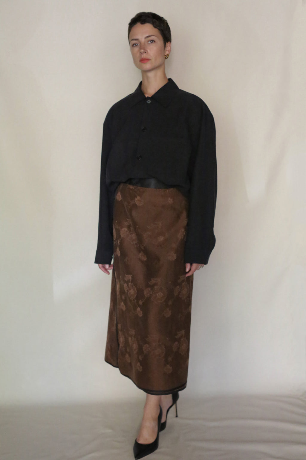 A person stands against a plain light background, wearing a loose black button-up shirt, the Lingering Fragrance Futo Skirt with a brown floral pattern, and black high-heeled shoes. They have short hair and a neutral expression.