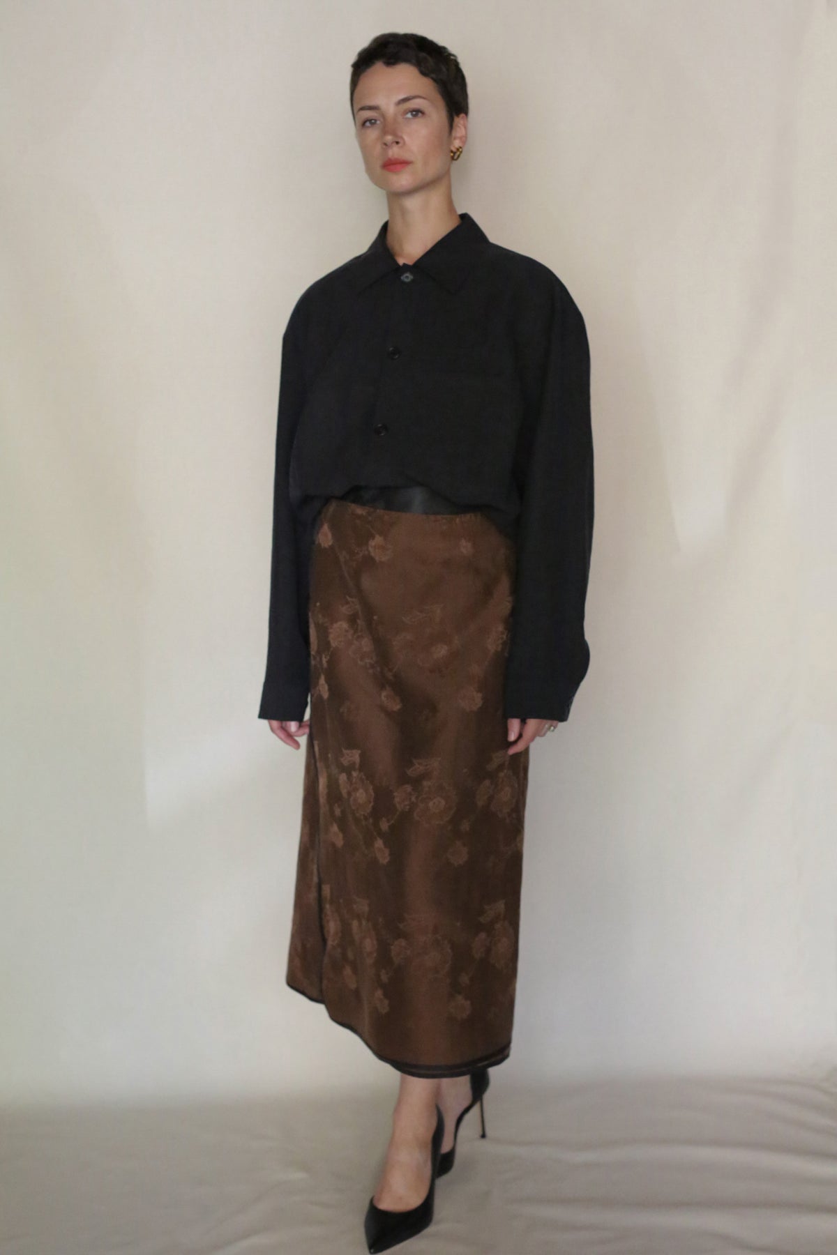 A person with short dark hair and a neutral expression stands against a plain background wearing the Earth Mark Men's Shirt in black, a brown floral-printed midi skirt dyed with plant-based dyes, and black pointed-toe heels.