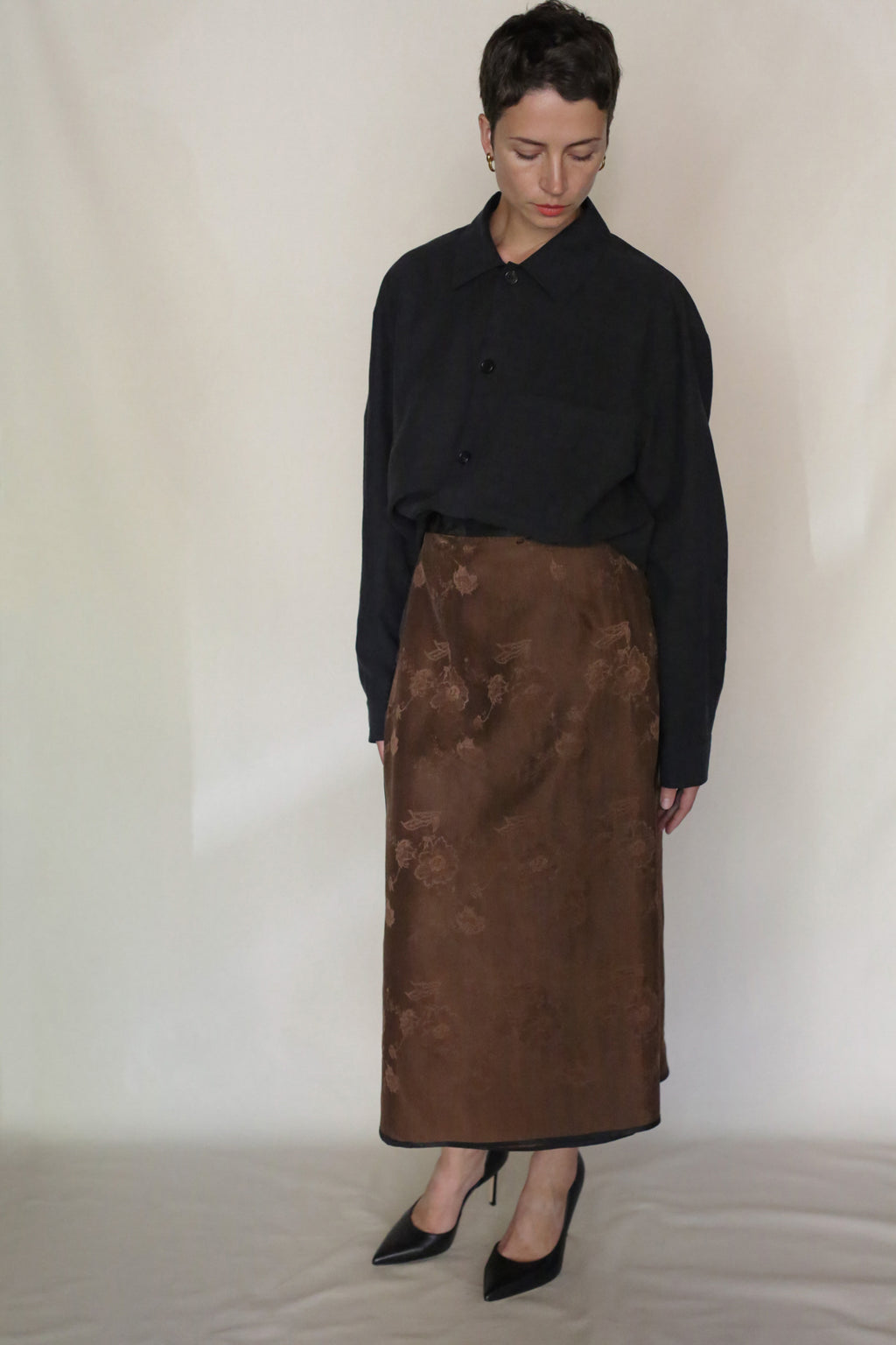 A person with short hair stands against a plain backdrop, wearing a loose black button-up shirt, black heels, and the Lingering Fragrance Futo Skirt—a brown silk jacquard floral midi skirt—while looking down.