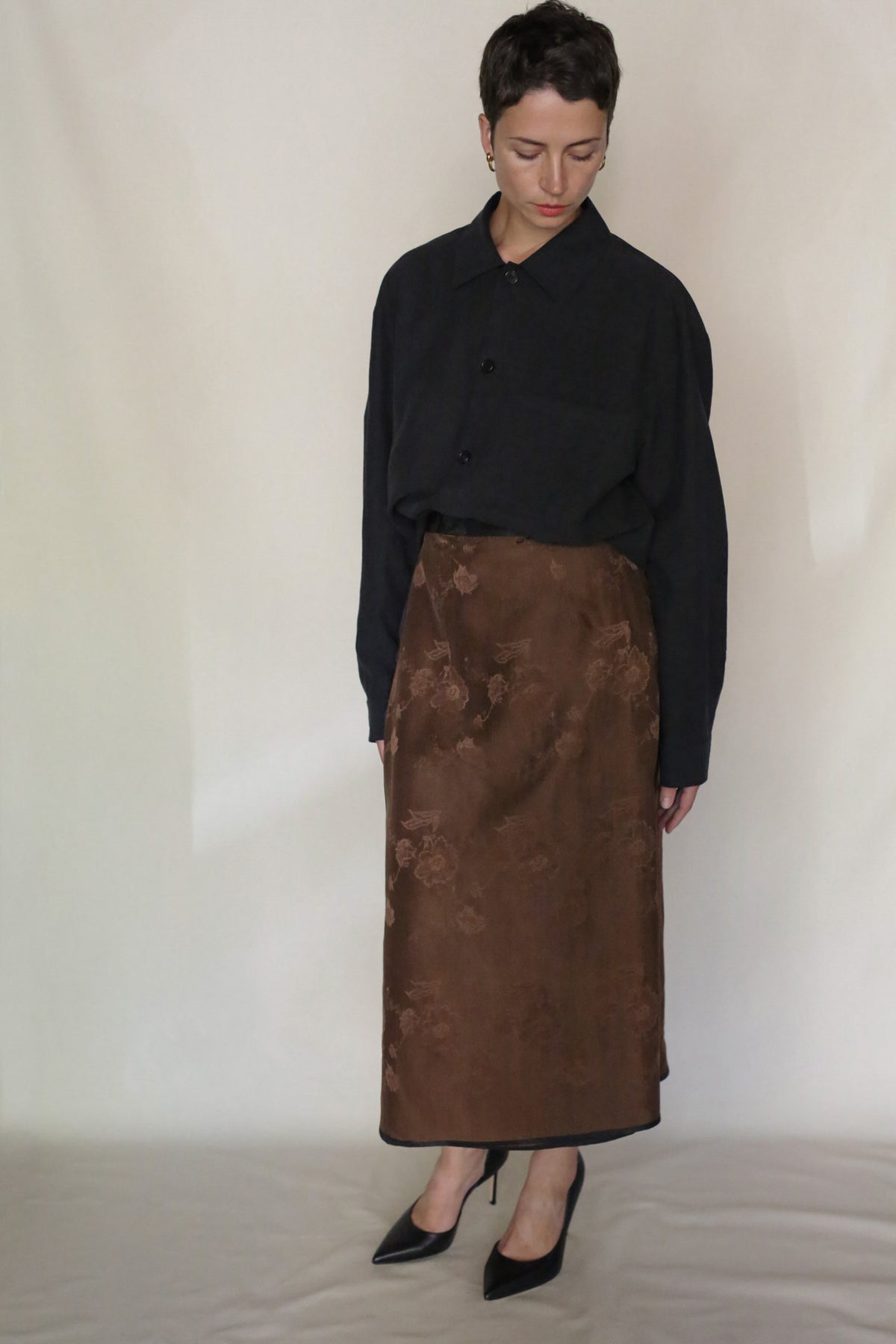 Wearing the Earth Mark Men's Shirt, a person with short hair pairs it with a brown floral ankle-length skirt and black high heels, standing against a plain light background and gazing downward.