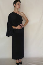 Load image into Gallery viewer, A person with short hair poses against a plain background, wearing the Eurie Dress in 2 Ply Silk Crepe Black and black heels. One arm is tucked behind their waist as they look slightly to the side.