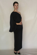 Load image into Gallery viewer, A person with short dark hair wears the Eurie Dress in 2 Ply Silk Crepe Black and black heels, standing confidently against a plain light background with hands at their waist—an elegant look embodying handmade San Francisco style.