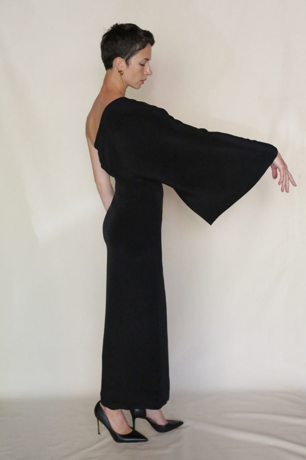 With short dark hair, a person stands sideways in the Eurie Dress—an asymmetrical, one-shoulder silk crepe design with a wide long sleeve and black heels—against a plain light background. This unique dress is handmade in San Francisco.