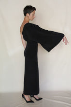 Load image into Gallery viewer, A person in the Eurie Dress | 2 Ply Silk Crepe Black stands sideways against a plain light background, one arm extended while the asymmetrical, handmade San Francisco gown drapes elegantly and pairs with black high heels.