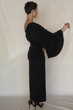 Load image into Gallery viewer, A person with short hair stands sideways against a plain light background, wearing the Eurie Dress in 2 Ply Silk Crepe Black with wide sleeves and black high heels, one arm bent and slightly raised.