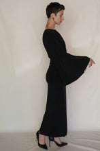 Load image into Gallery viewer, A person with short dark hair, wearing the Eurie Dress | 2 Ply Silk Crepe Black and black high heels, stands in profile against a plain light background, pointing downward with their right hand.
