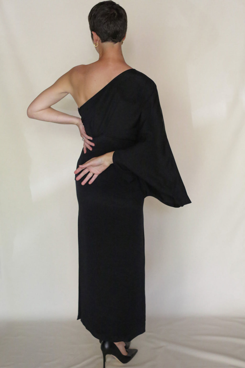 Wearing the Eurie Dress, a person in a long, black, asymmetrical one-shoulder gown and black heels stands facing away with one hand on their lower back and the other bent across, set against a plain light background.