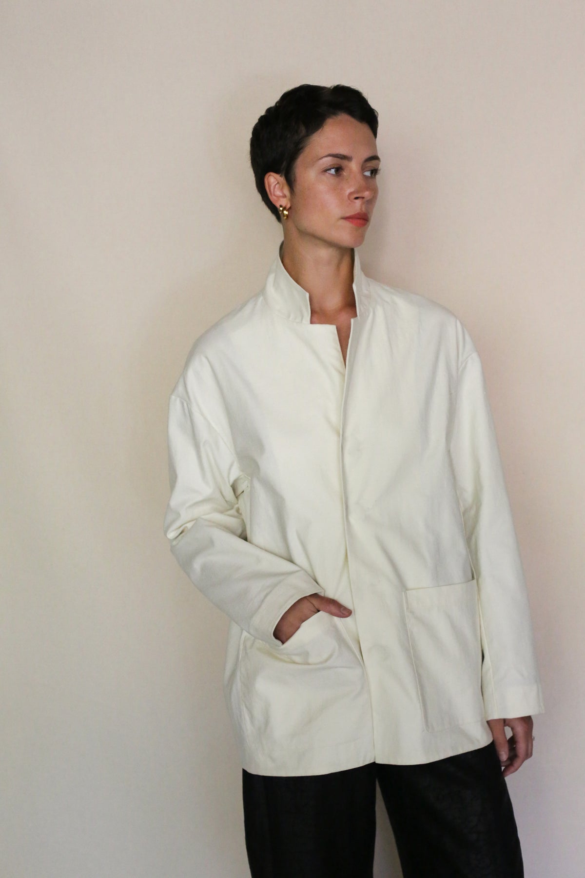 A person with short dark hair wears the Painter's Jacket | Undyed—handmade in San Francisco from cotton serge with large pockets—paired with black pants, standing against a plain background and looking to the side with one hand in their pocket.