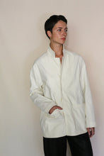 Load image into Gallery viewer, A person with short dark hair, wearing the Painter's Jacket | Cotton Serge Ivory and black pants, stands against a beige background, gazing to the side with one hand in their pocket. The handmade San Francisco style defines their look.