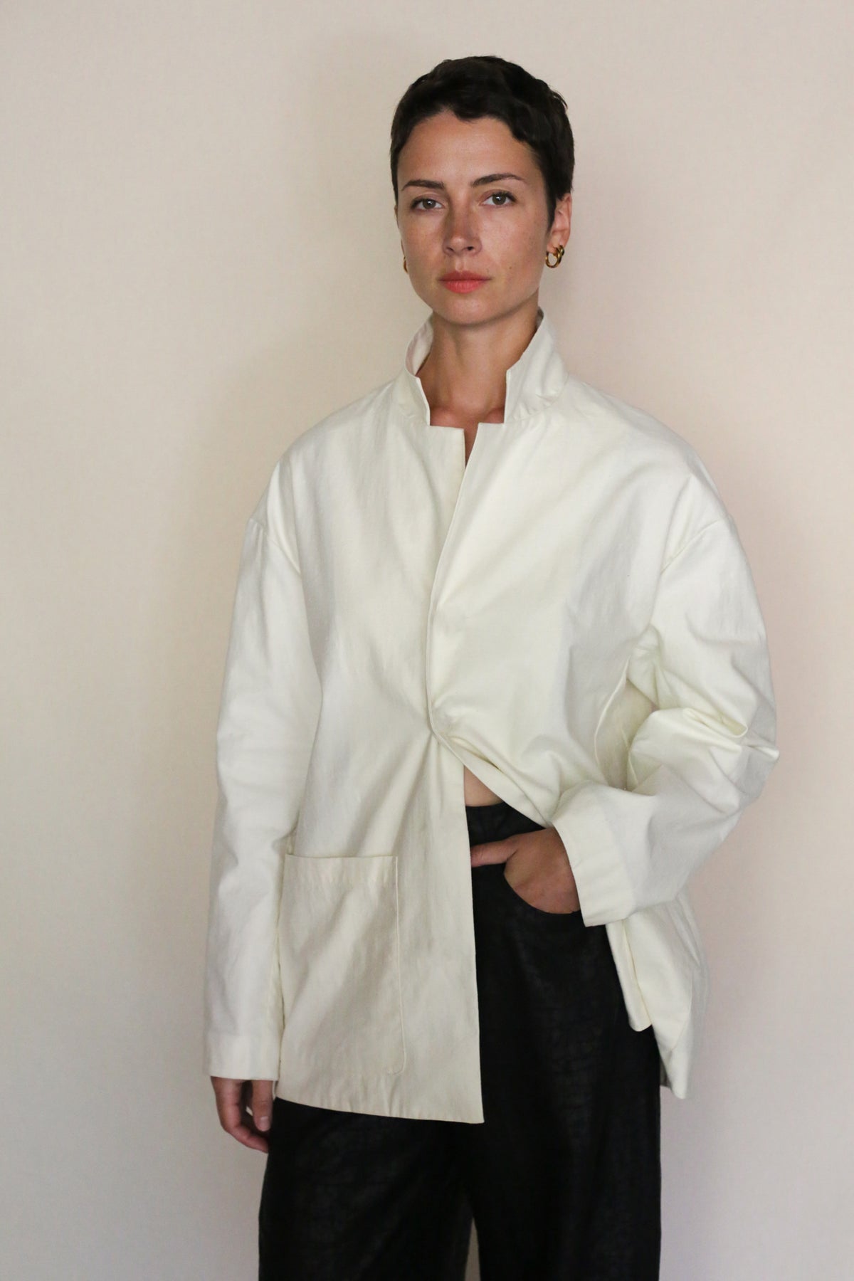 A person with short dark hair wears the handmade Painter's Jacket | Undyed, crafted from cotton serge, paired with black pants. They stand against a plain light background, one hand in their pocket. Made in San Francisco.