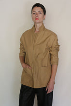 Load image into Gallery viewer, A person with short dark hair wears the Painter's Jacket | Cotton Serge Tan, featuring pockets, paired with black pants. They stand against a light background, looking confidently at the camera with one hand in a pocket.