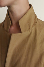 Load image into Gallery viewer, A close-up of a person wearing the Painter's Jacket | Cotton Serge Tan, handmade with light brown cotton serge and a mandarin collar, showing their neck and jawline against a plain background.