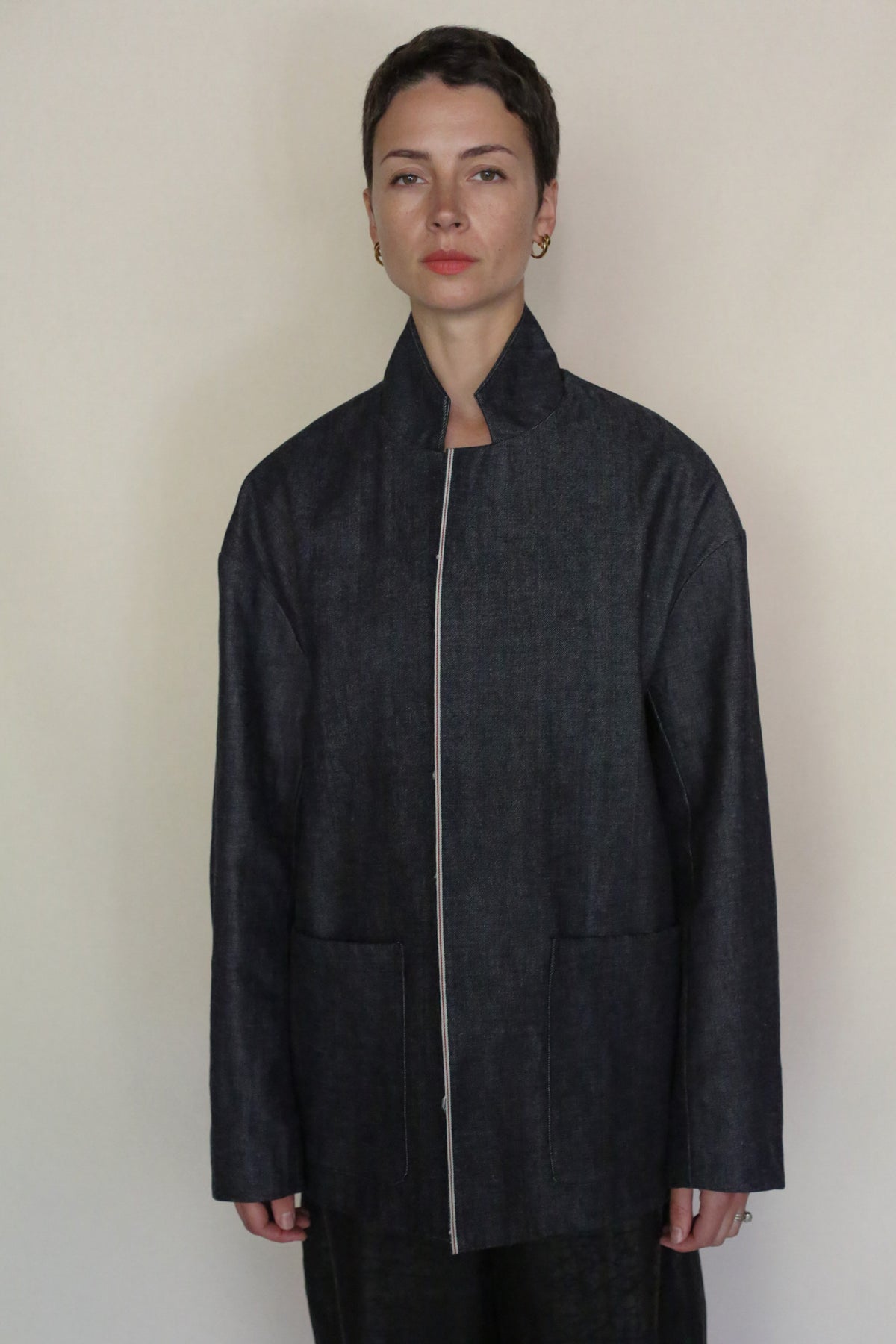 A person with short hair stands facing forward, wearing the Painter's Jacket | Japanese Selvedge Denim—a dark, minimalist jacket with a high collar and large front pockets. The background is plain and light-colored.