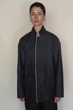 Load image into Gallery viewer, A person with short hair stands facing forward, wearing the Painter's Jacket | Japanese Selvedge Denim—a dark, minimalist jacket with a high collar and large front pockets. The background is plain and light-colored.