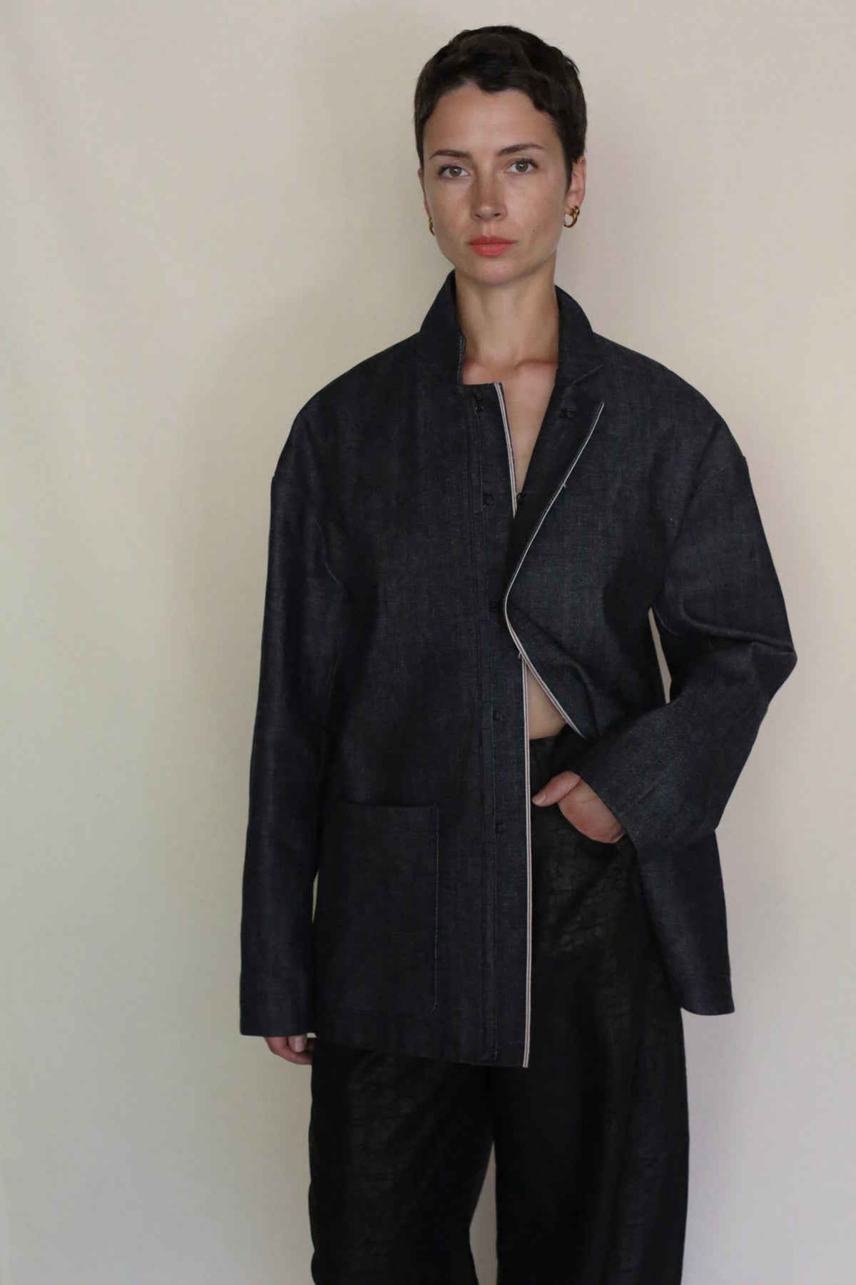 A person with short dark hair stands against a plain background, wearing the Painter's Jacket | Japanese Selvedge Denim over matching wide-leg pants, one hand in a pocket and a calm expression.
