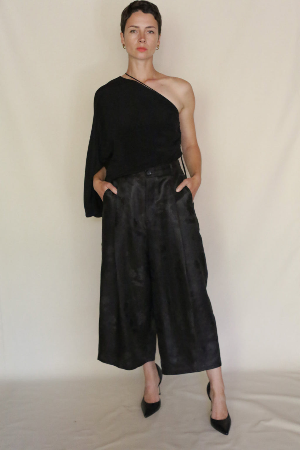 A person poses confidently against a plain backdrop, wearing Samurai Pants and a black one-sleeve top. The sculptural, cropped silhouette is paired with black heels, hands in pockets for a bold look.