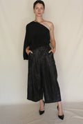 A person poses confidently against a plain backdrop, wearing Samurai Pants and a black one-sleeve top. The sculptural, cropped silhouette is paired with black heels, hands in pockets for a bold look.