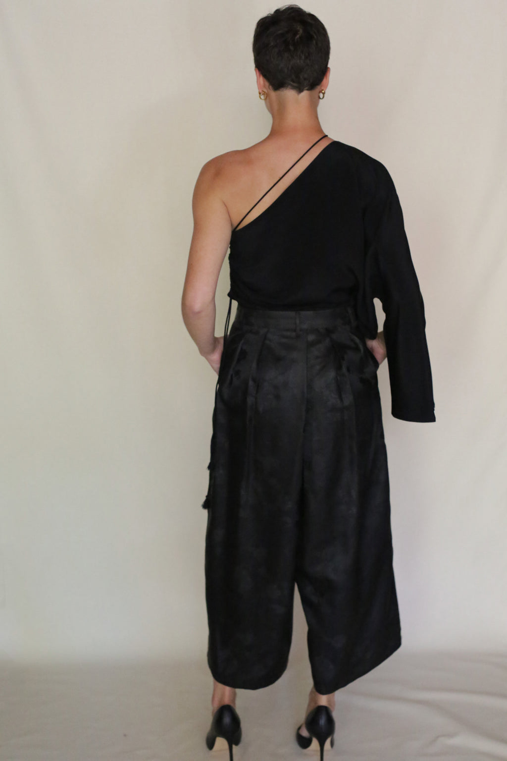 A person with short hair, seen from behind, wears Samurai Pants and a black one-shoulder cropped top with one sleeve off the shoulder, exposing the back and one arm, paired with black heeled shoes.